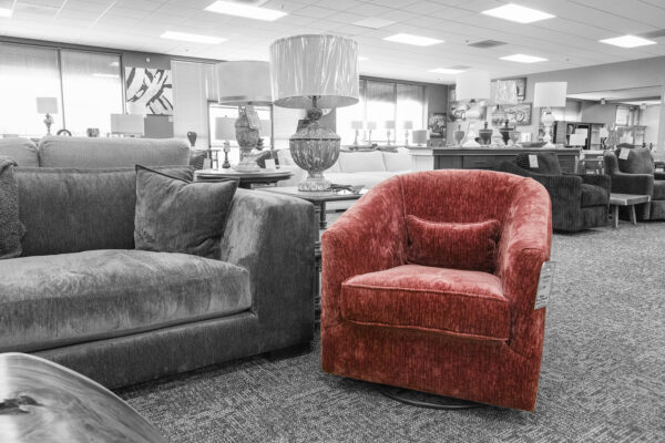 Red barrel swivel chair with a matching lumbar pillow, positioned in a showroom next to a gray sofa and table lamps.