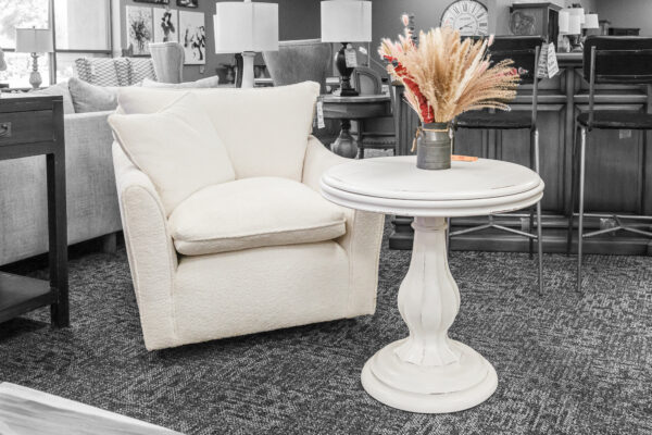 White upholstered lounge chair with a matching round pedestal table, set against a backdrop of layered living room displays.
