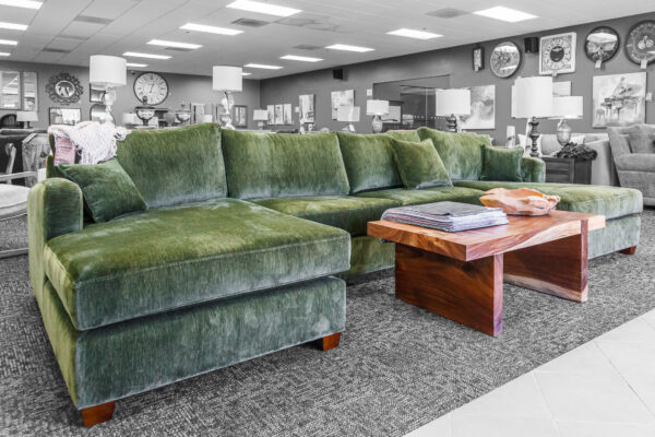 Oversized green velvet sectional with a wide chaise, paired with a modern live-edge coffee table in a furniture showroom.
